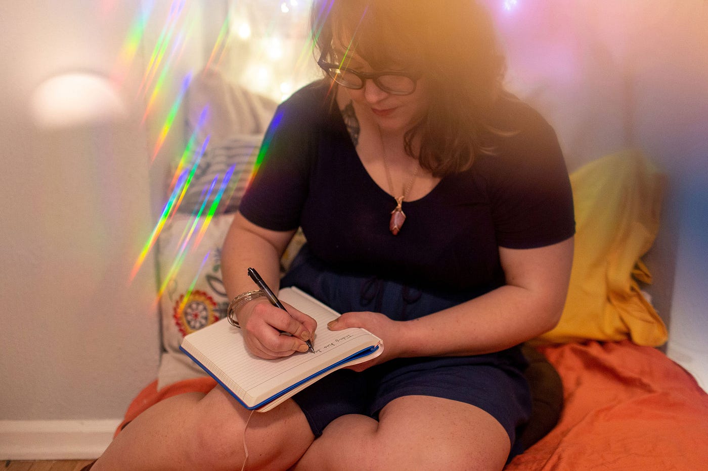 Photo of Allison writing in her journal. She is wearing a dark colored outfit and crystal necklace while sitting in a cozy nook of pillows. There is a photography effect used that gives the photo a blurred quality in spots and casts lines of rainbow prisms in others.