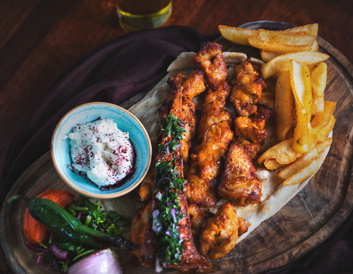 Grilled chicken kebab, chips, spiced yoghurt and garnish served on a chopping board.
