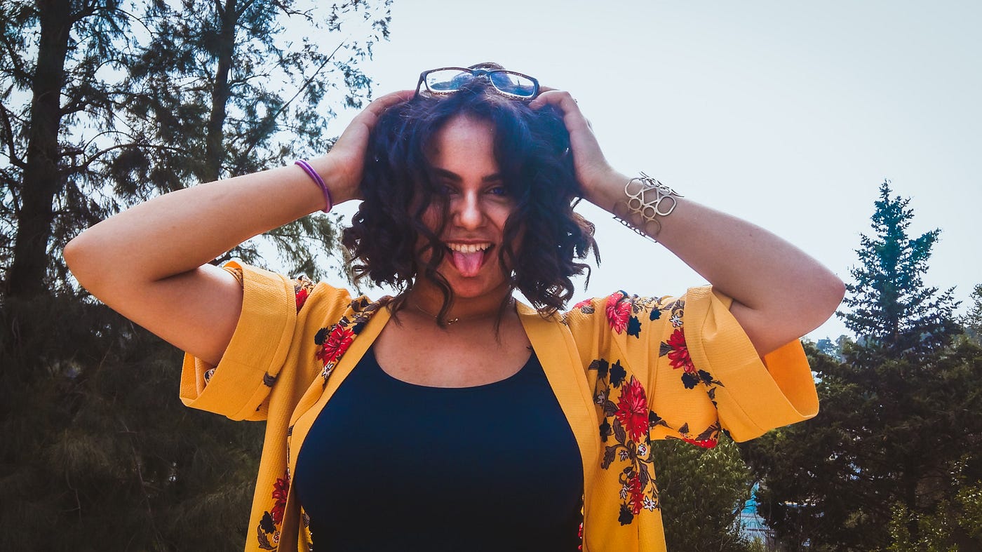 woman looking confused tongue out holding top of her head wearing bright yellow and black