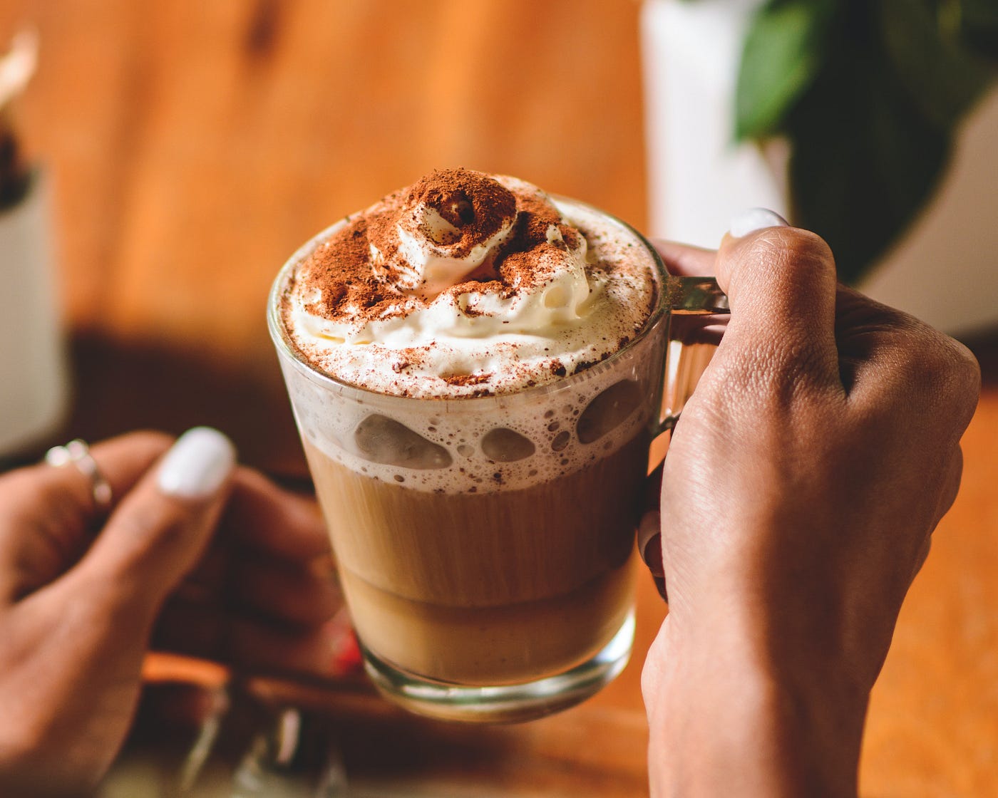 Brown hands holding a clear mug of hot chocolate with whipped cream