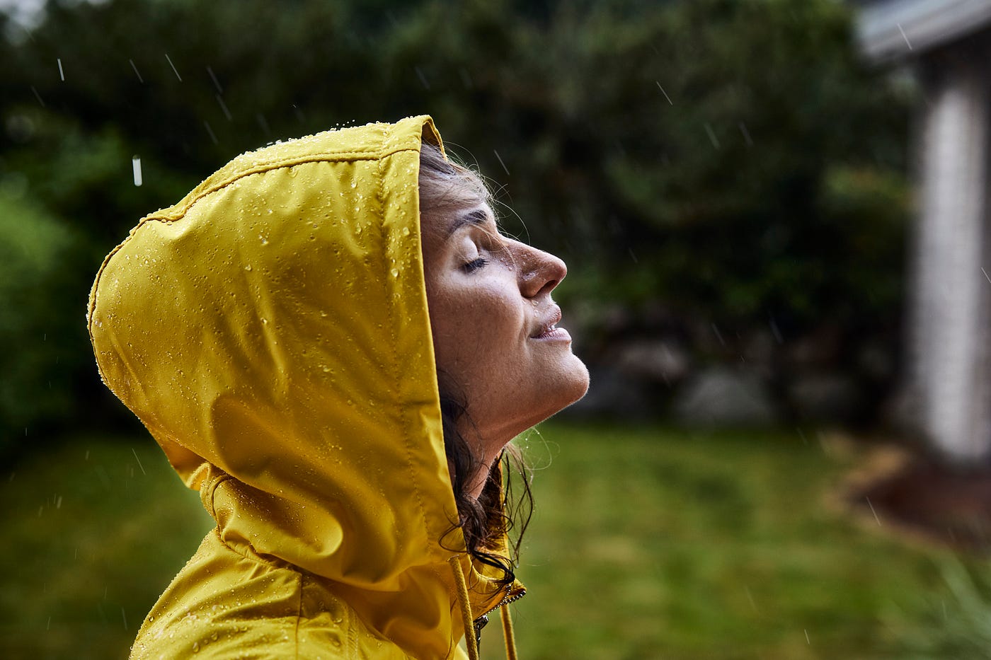 A photo of a woman wearing a raincoat and hood. She is standing in the rain with her eyes closed, looking peaceful.