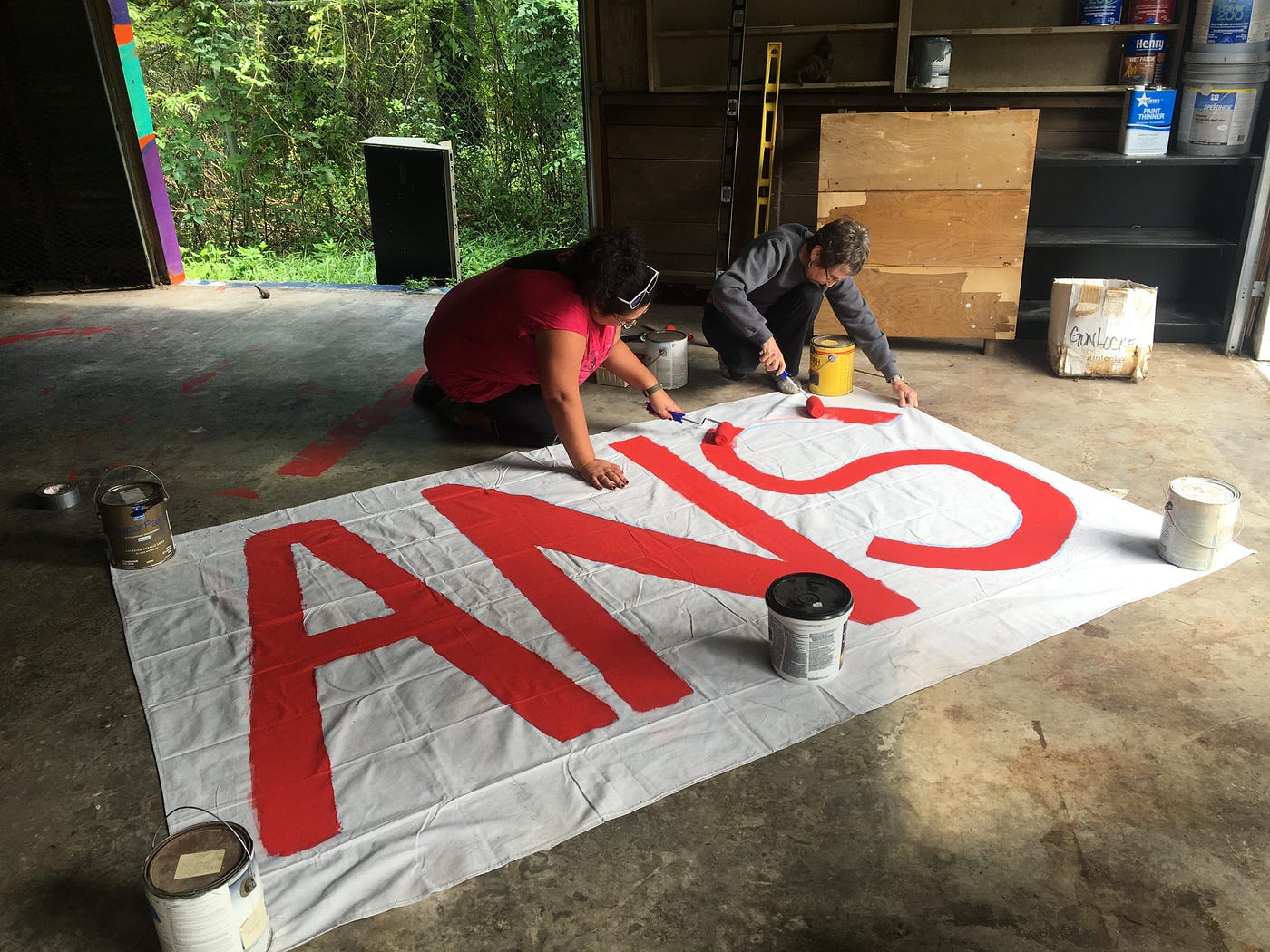 Two volunteers paint a canvas with large red letters on a garage floor.