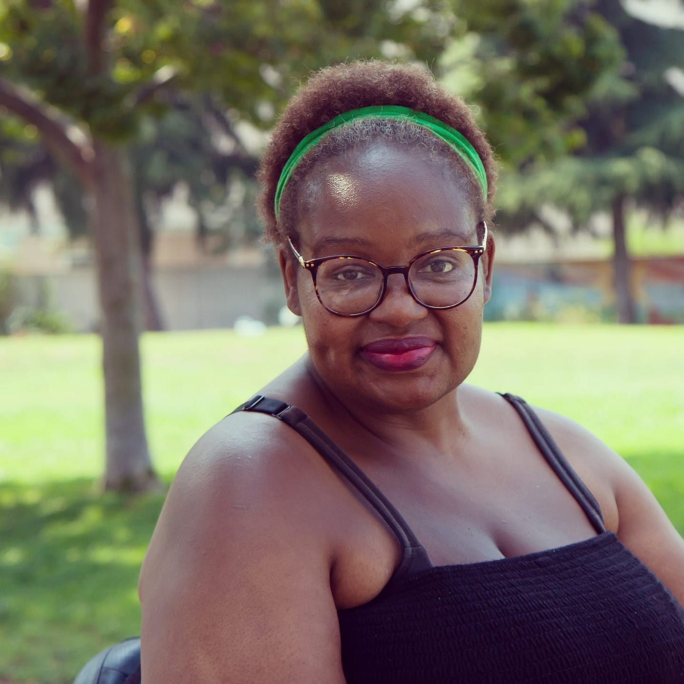 A Black woman with short hair, a green headband, brown-framed glasses, a black tank top & pink lipstick smiles cutely.