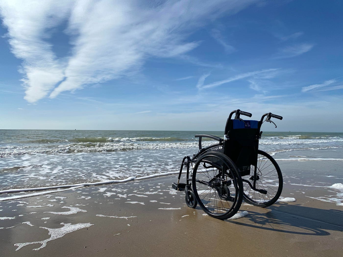 A wheelchair by the ocean; waves are lapping up the beach under a blue sky