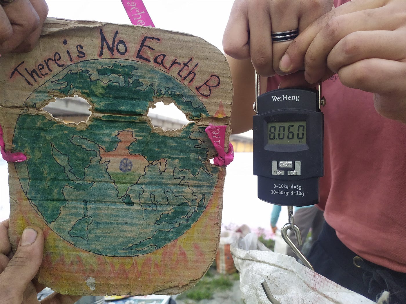 A person holds a weighing machine which indicates the weight of the bag they’re holding is 8.060kg while another person holds a small board beside him which says: ‘There Is No Earth B’