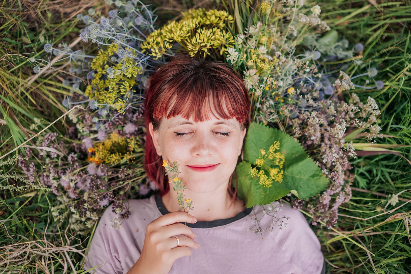 A young, redhead, freckled woman with a peaceful smile laying in a pile of flowers, eyes closed and a flower in hand.