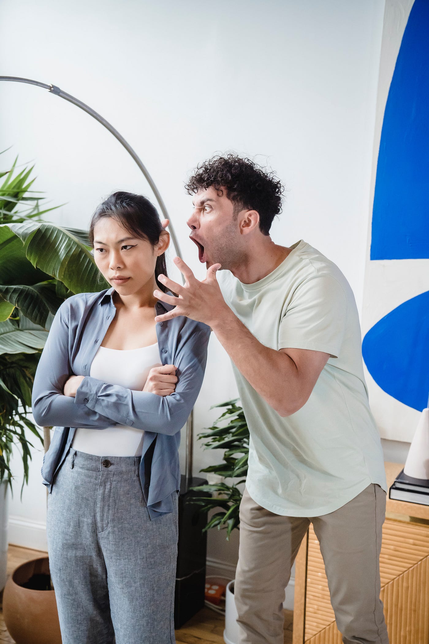 A man with curly hair shouts aggressively in the ear of a woman, whose back is turned toward him, with her arms crossed.