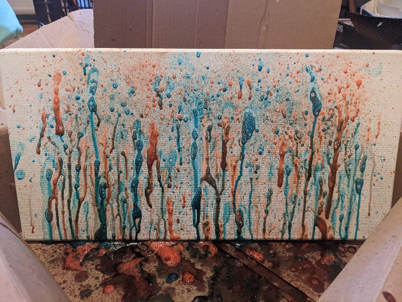A canvas is placed in a cardboard box with a cardboard screen behind it. On the canvase is blue and red paint dripping down, imitating a forest.