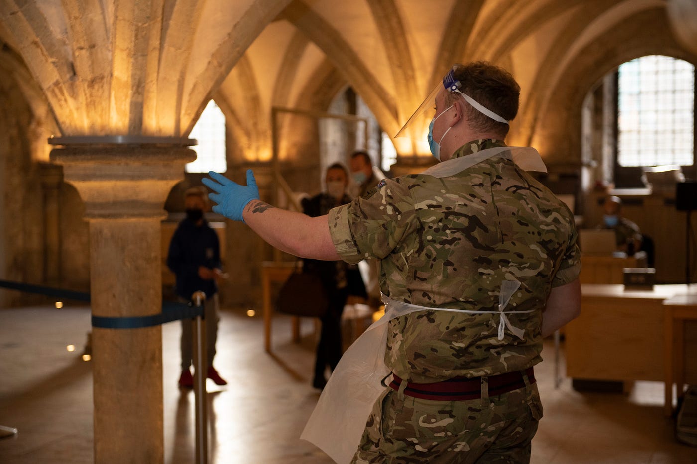 Under a vaulted ceiling, a soldier in PPE with his back to the camera directs members of the public