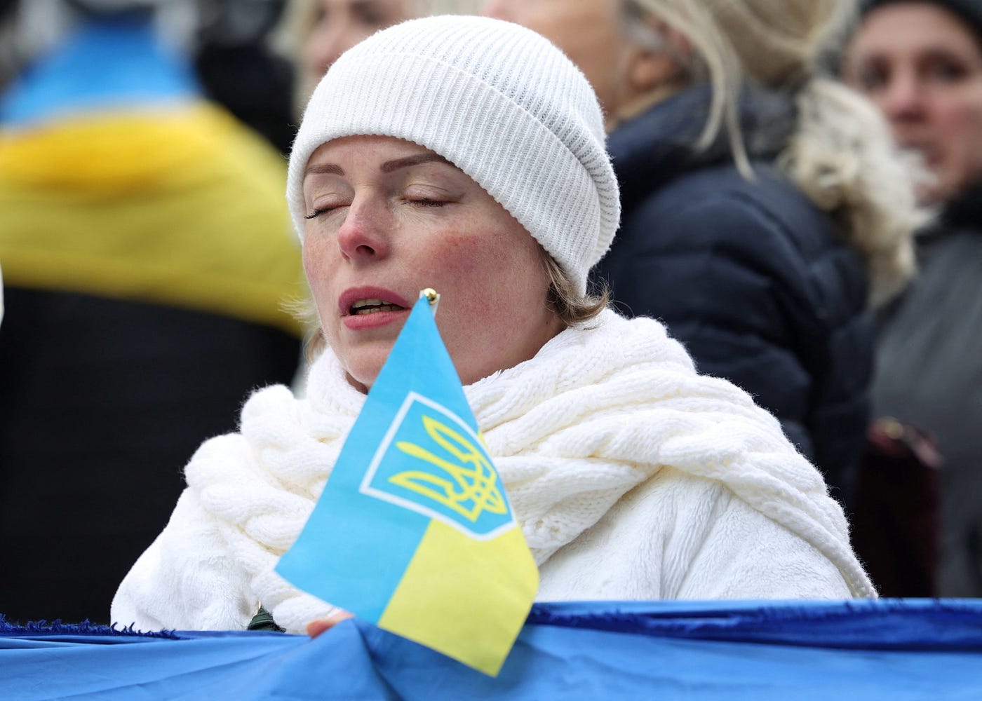Woman in wnite wool cap and winter clothng stands outdoors with eyes closed in prayer and holds the Ukrainian flag in one of the worldwide demonstrations seeking and end to the Russian invasion of Ukraine.