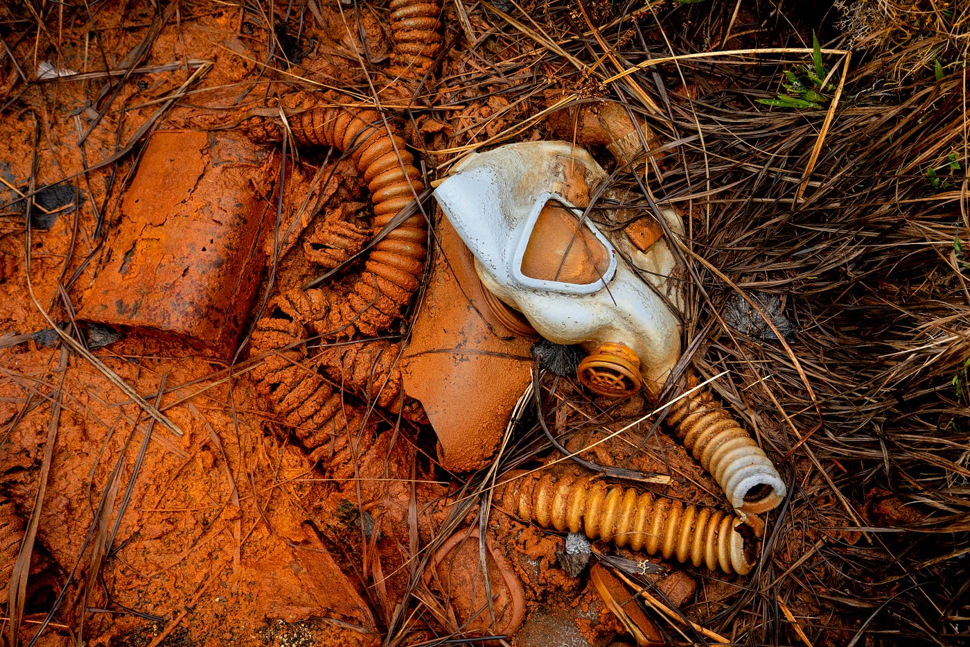 A silver metal face shield lies in rust-colored mud, tubing and other debris is half submerged in the mud, and grass shows at the edges.