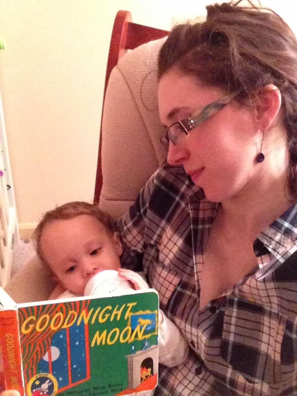 Annie sits in a chair holding Emmy as a baby as she drinks a bottle. Annie is looking at Emmy and Emmy is looking at the book Goodnight Moon.