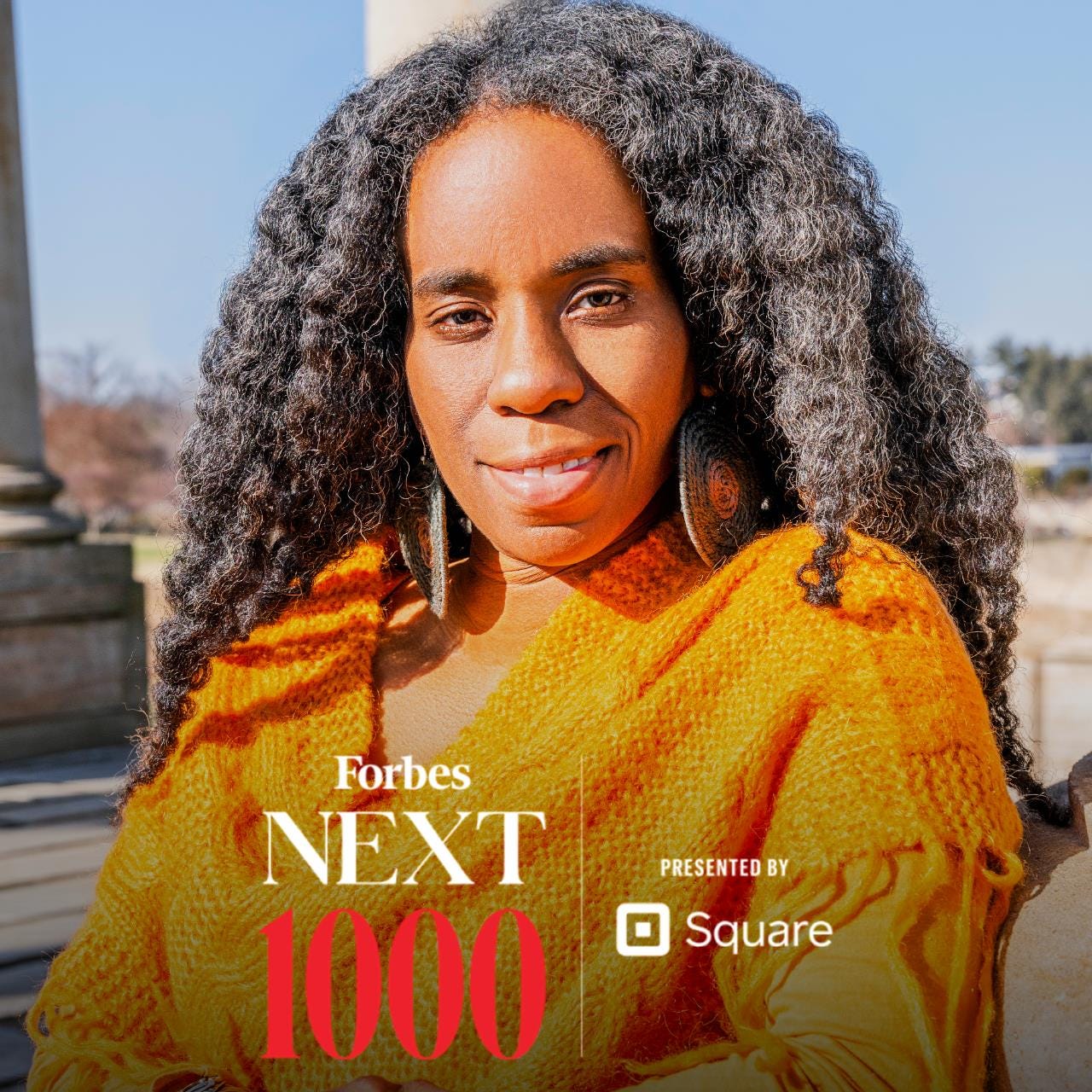 Alyscia Cunningham with graphic that reads “Forbes Next 1000”. She is outdoors and wears a mustard yellow sweater throw. She smiles gently staring directly into the camera. Her hair is black and is out and wavey.