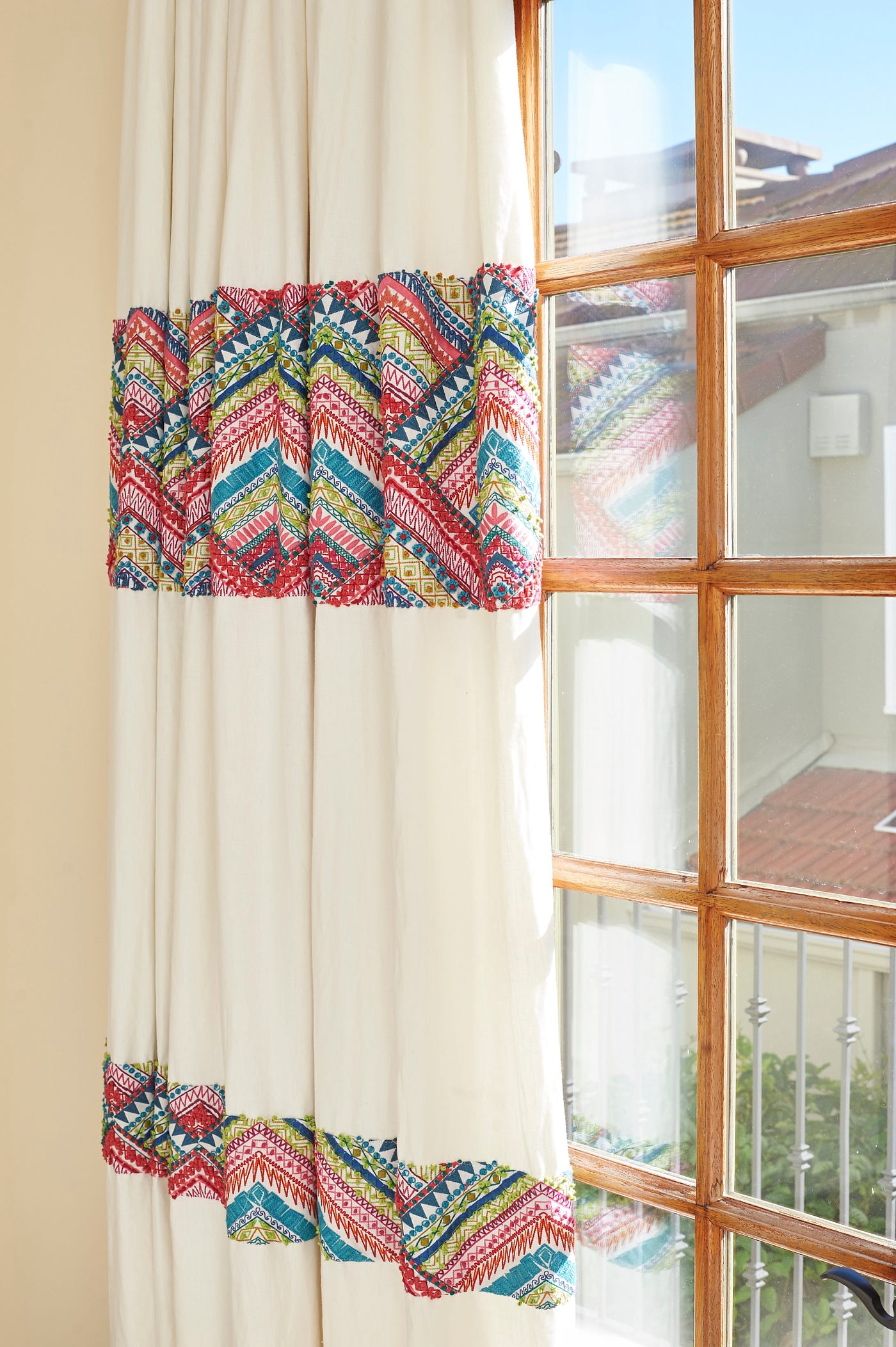 Handmade creamy-white draperies banded with colorful embroidery in shades of fuchsia, teal, chartreuse and gold hanging against a paned window.