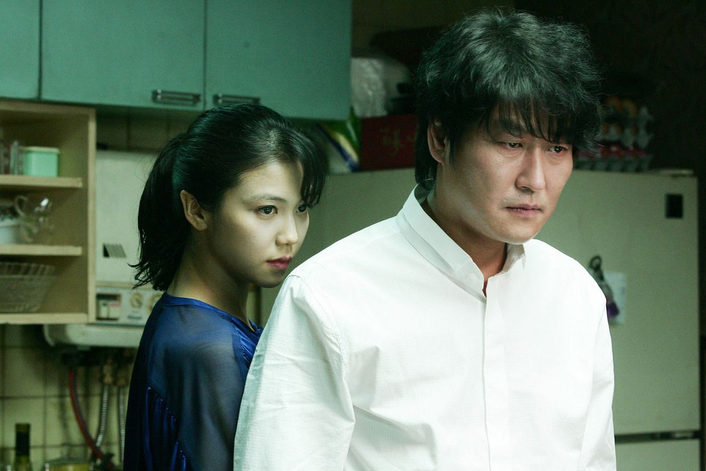 A woman is seen standing behind a man in a kitchen. They are both staring in the same direction.