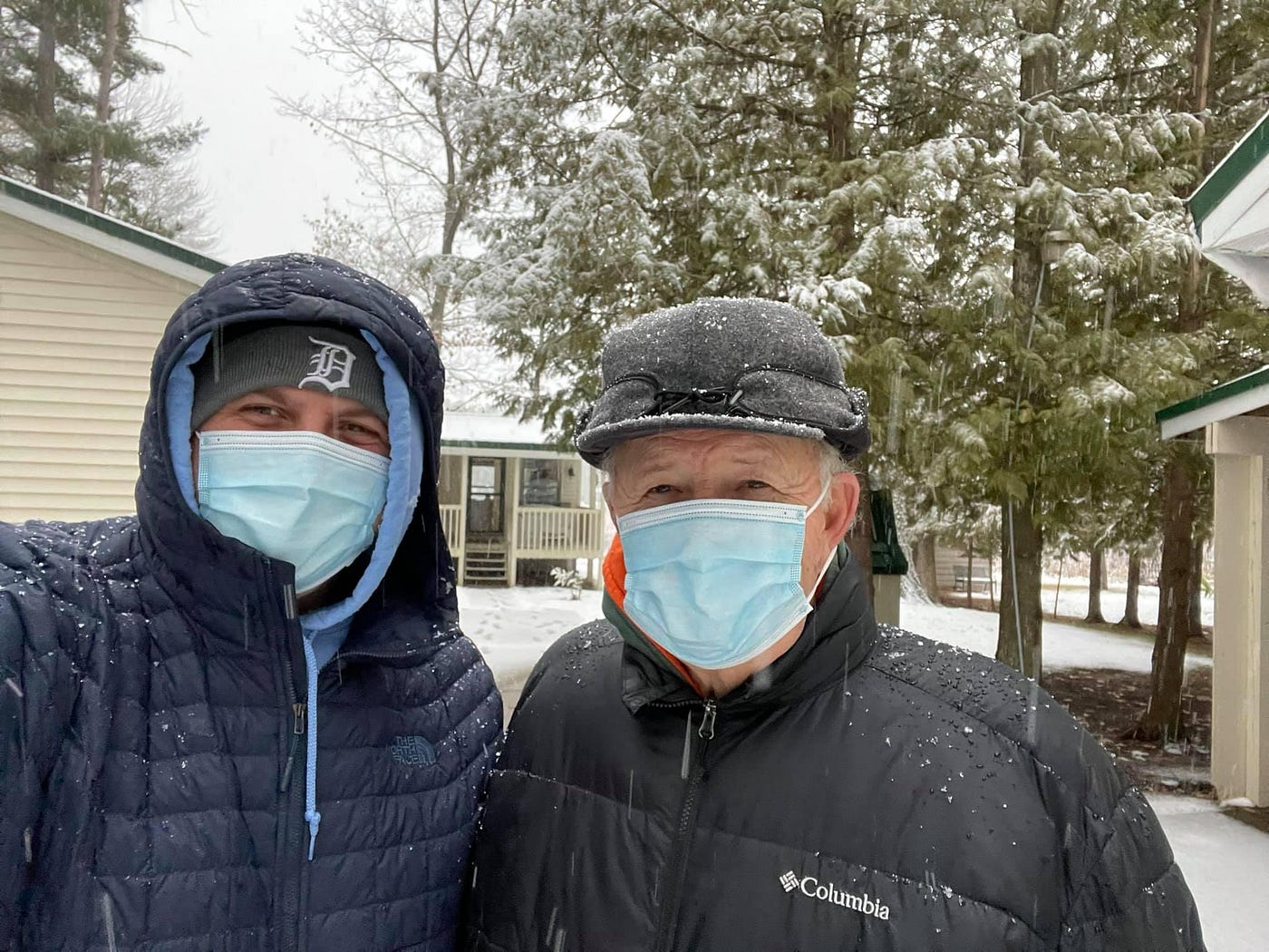 Father and son stand in front of the cabin while it’s snowing dressed in winter coats, hats, and surgical masks.