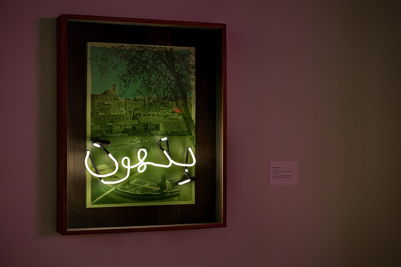 A green-tinted photograph of a city on the edge of the water is framed in a large wood frame. A neon sign reading Bit-hoon (this too shall pass) is installed over the background image.
