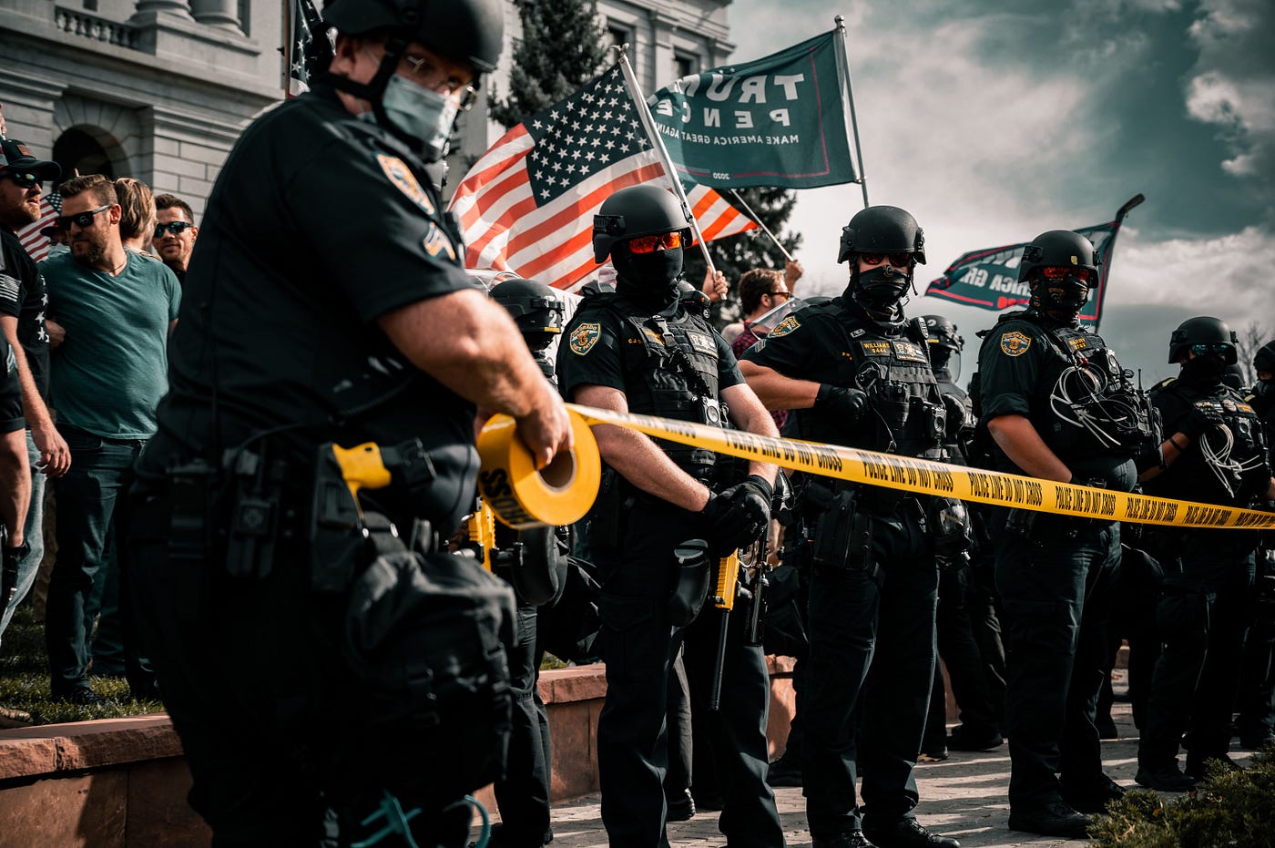 Police unrolling yellow tape in front of a crowd holding Trump/Pence and USA flags
