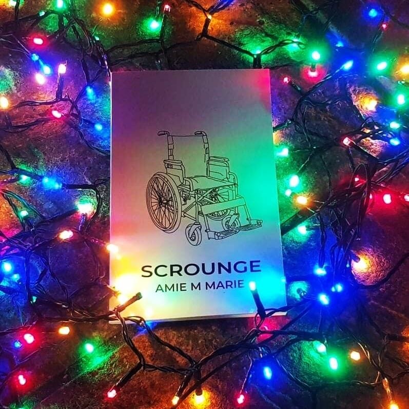 A physical copy of the book of the play ‘Scrounge’ by Amie M Marie. The book rests on top of a christmas tree, lit by a bright constellation of fairy lights in different colors.