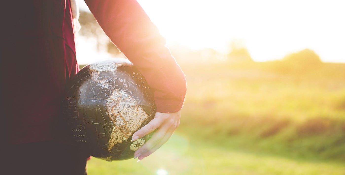 The back of a woman in a red shirt, holding a globe against her body with one hand as she looks out into bright sunlight