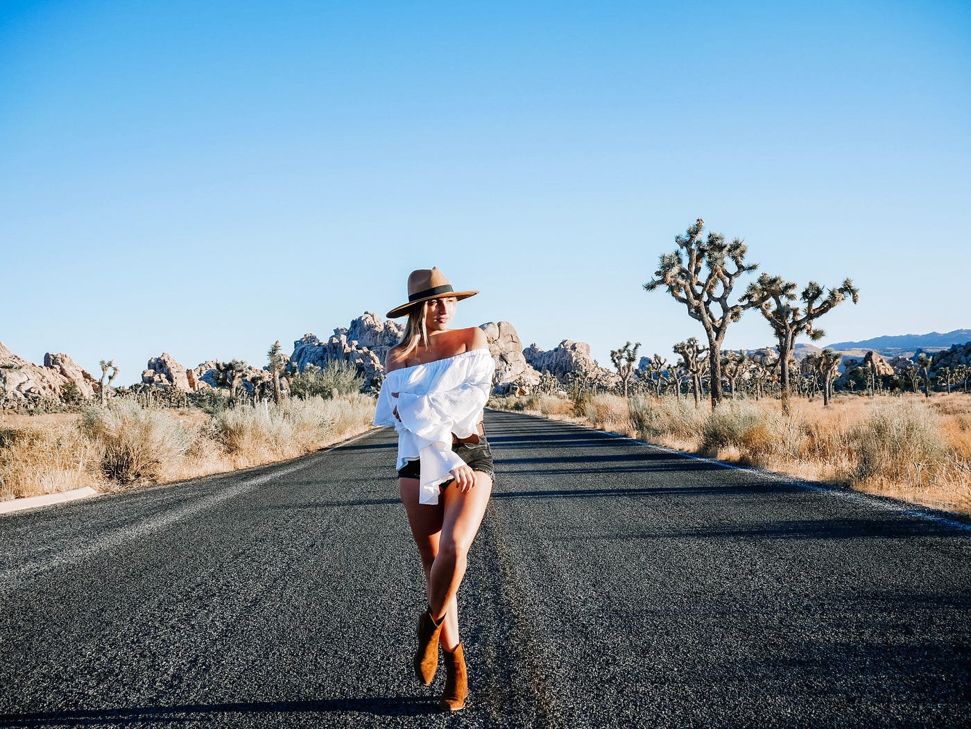 White woman in white shoulderless top and shorts, brimmed hat and boots, standing in the middle of a road in Joshua Tree Park