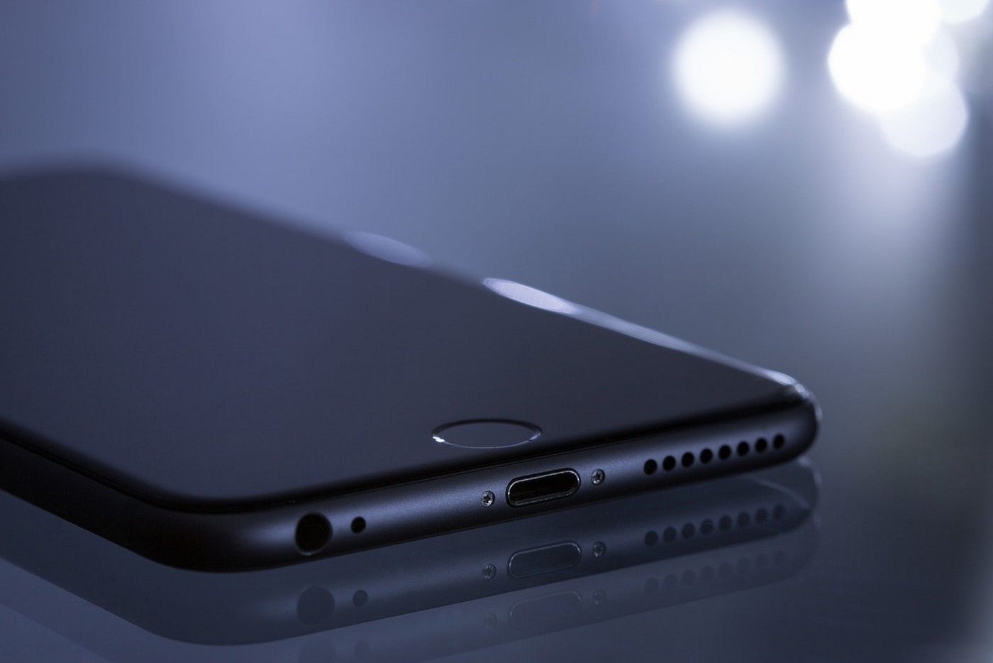 Color photo of a black mobile phone, face down on a surface. Bright lights in the background of otherwise darkness.