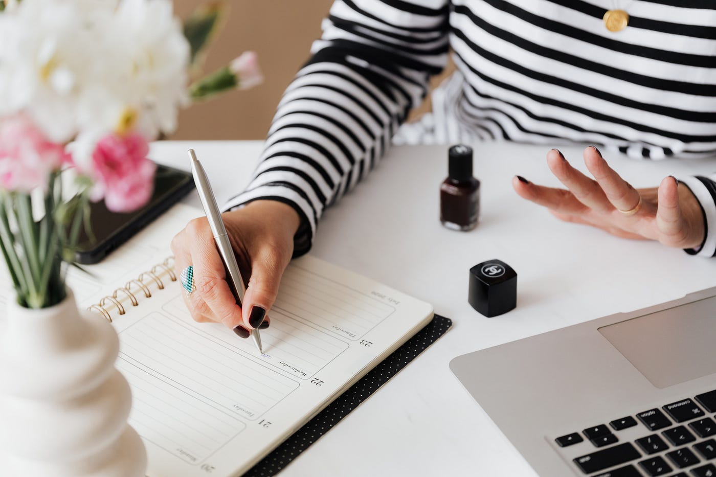 An individual writing in a planner on a neat white desk, with flowers, nail polish, and a laptop. Individual is wearing a white and black striped shirt.