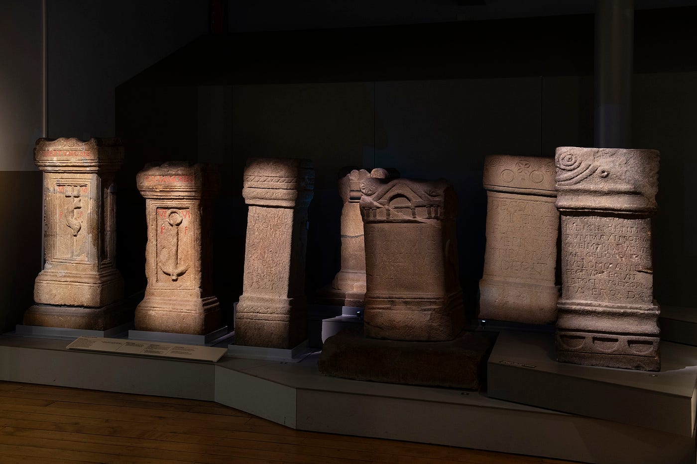 Seven Roman sandstone altars in a museum. They are upright cuboid sculptures carved with Latin inscriptions and decorations.
