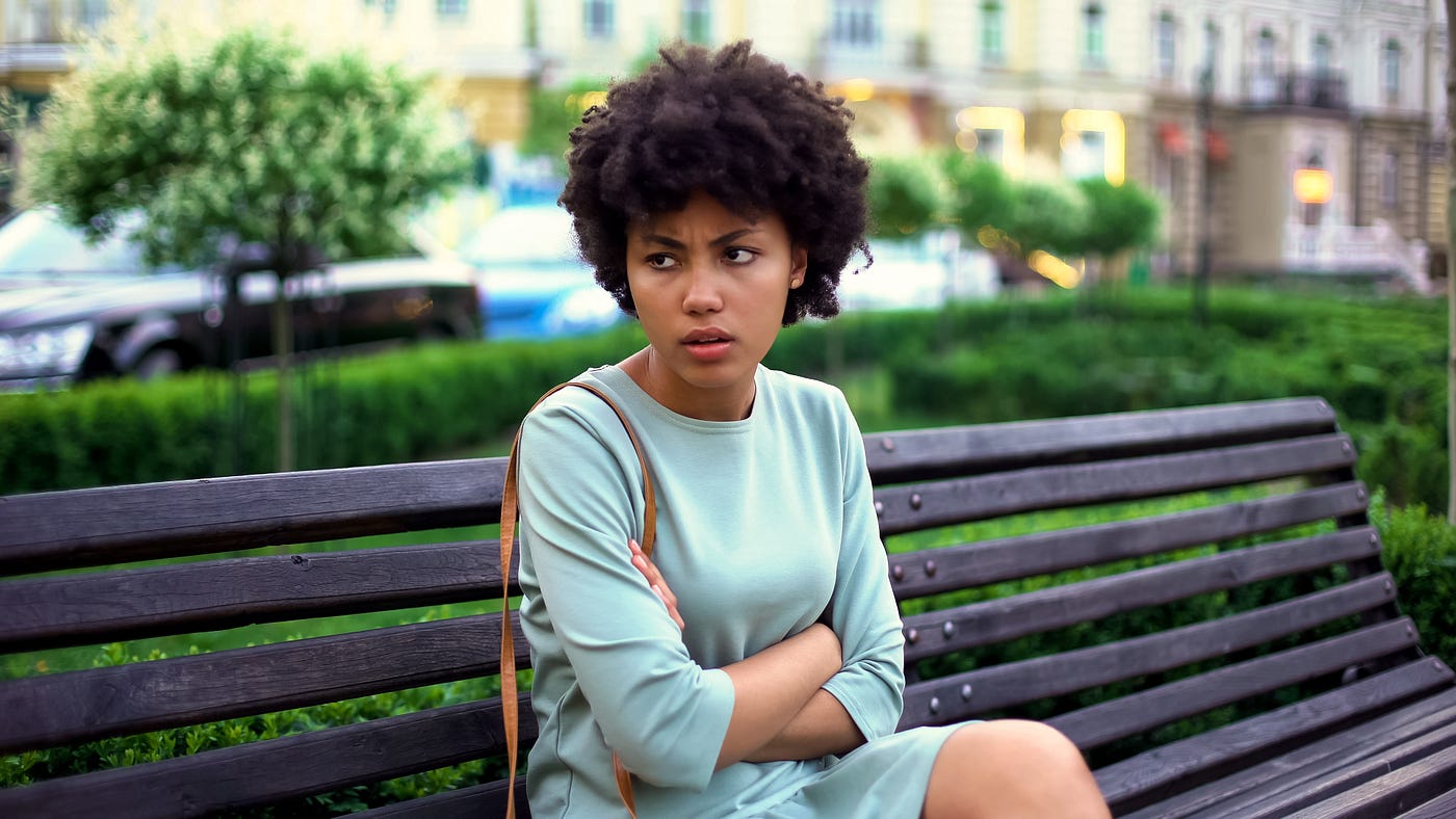 A photo of an annoyed black woman sitting on a park bench.