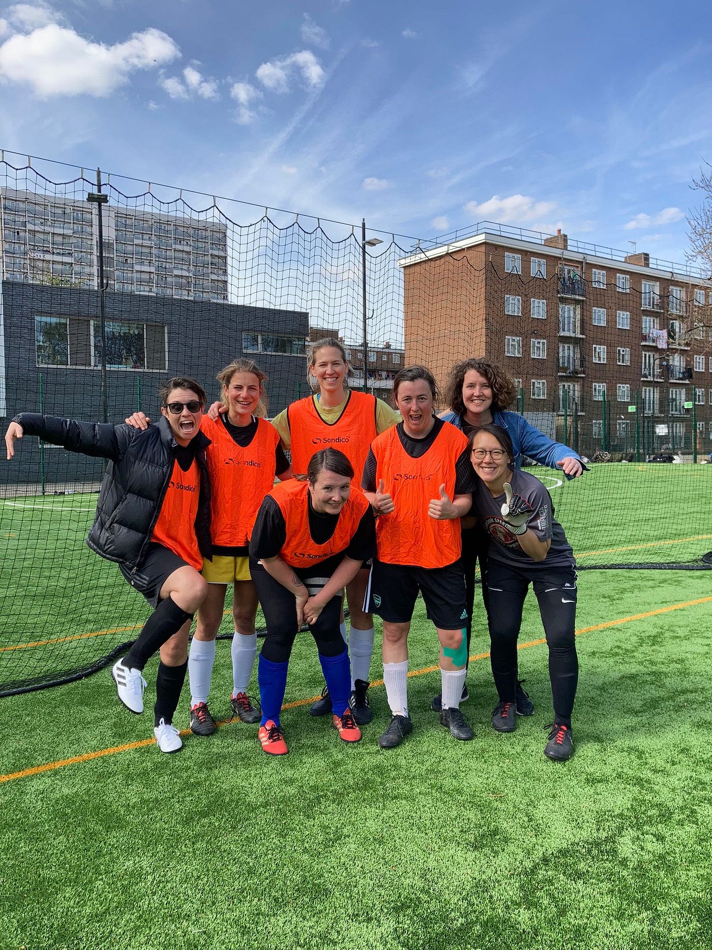 A football team wearing orange bids standing in front of a black net on top of astro turf. They are huddled together looking very happy