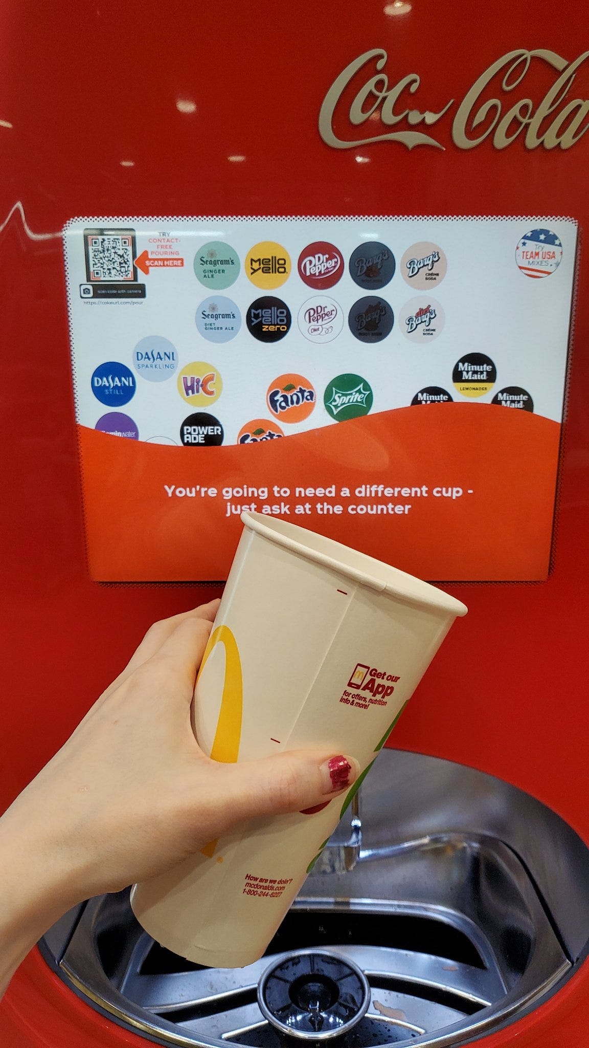 A self-service Coca Cola drink-dispensing machine displaying an error message: ‘You’re going to need a different cup. Just ask at the counter.’ A woman’s hand is holding a disposable wax-paper McDonald’s cup before the machine. Image: @AnemoneAndMe https://twitter.com/AnemoneAndMe/status/1420540789734612992