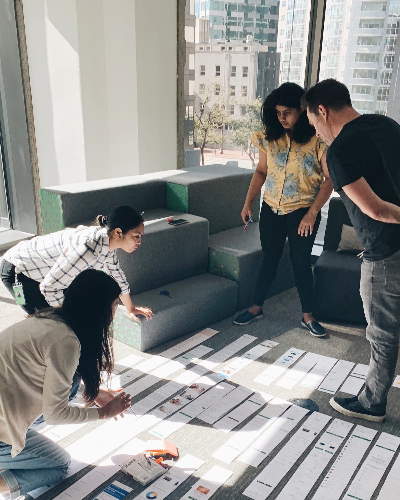 A photo of two people standing and two people kneeling. They’re all looking at screenshots of a website that are printed out in long strips of paper and lined up on the ground.