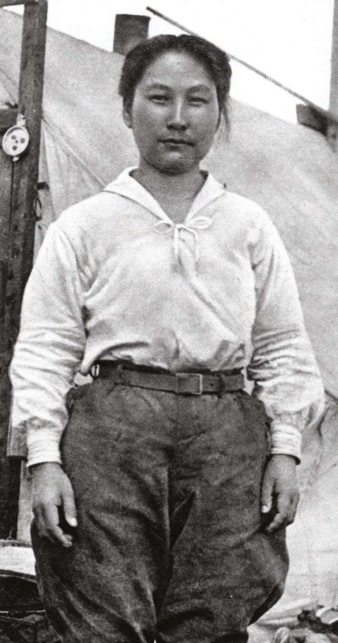 A woman stands in front of a tent in a black and white photo in a shirt and pants