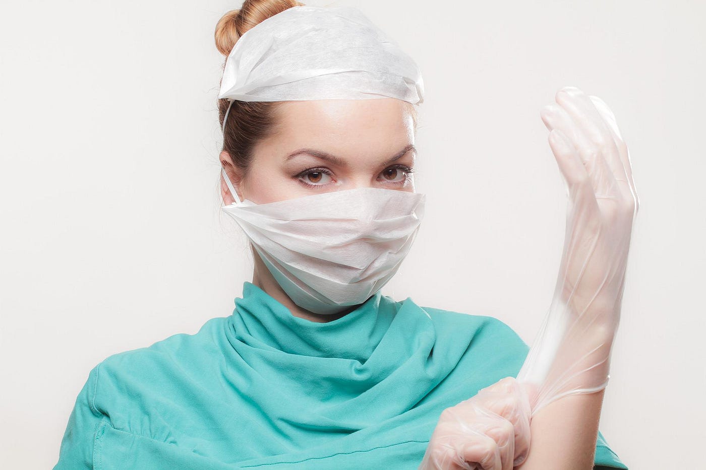 A dark-haired white female doctor glances meaningfully at us. She’s wearing turquoise protective clothing and two white masks. She’s just finished adjusting the fit of a transparent white plastic glove on her left hand.