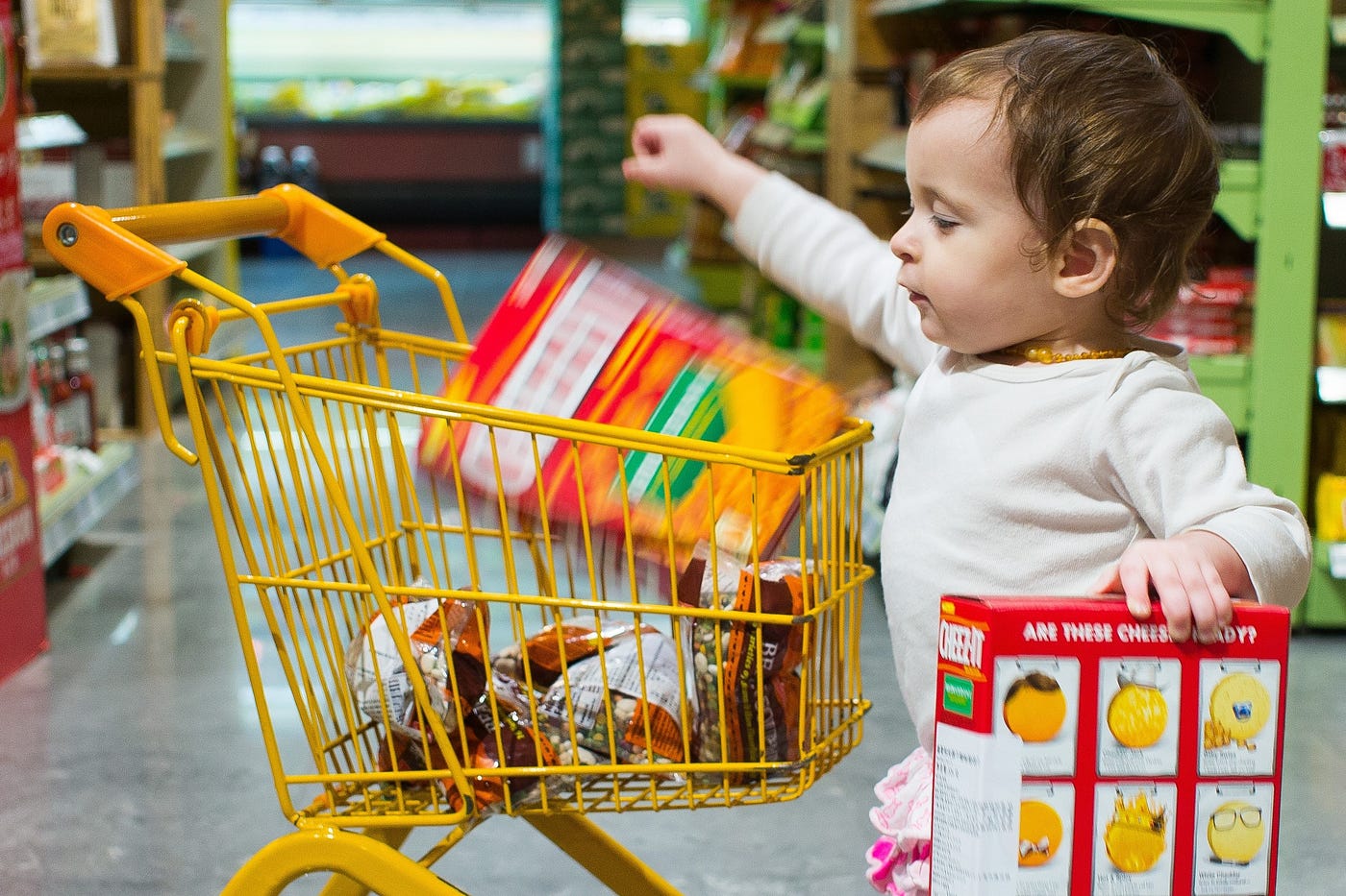 Toddler drops box of crackers into a small yellow shopping cart in a grocery store