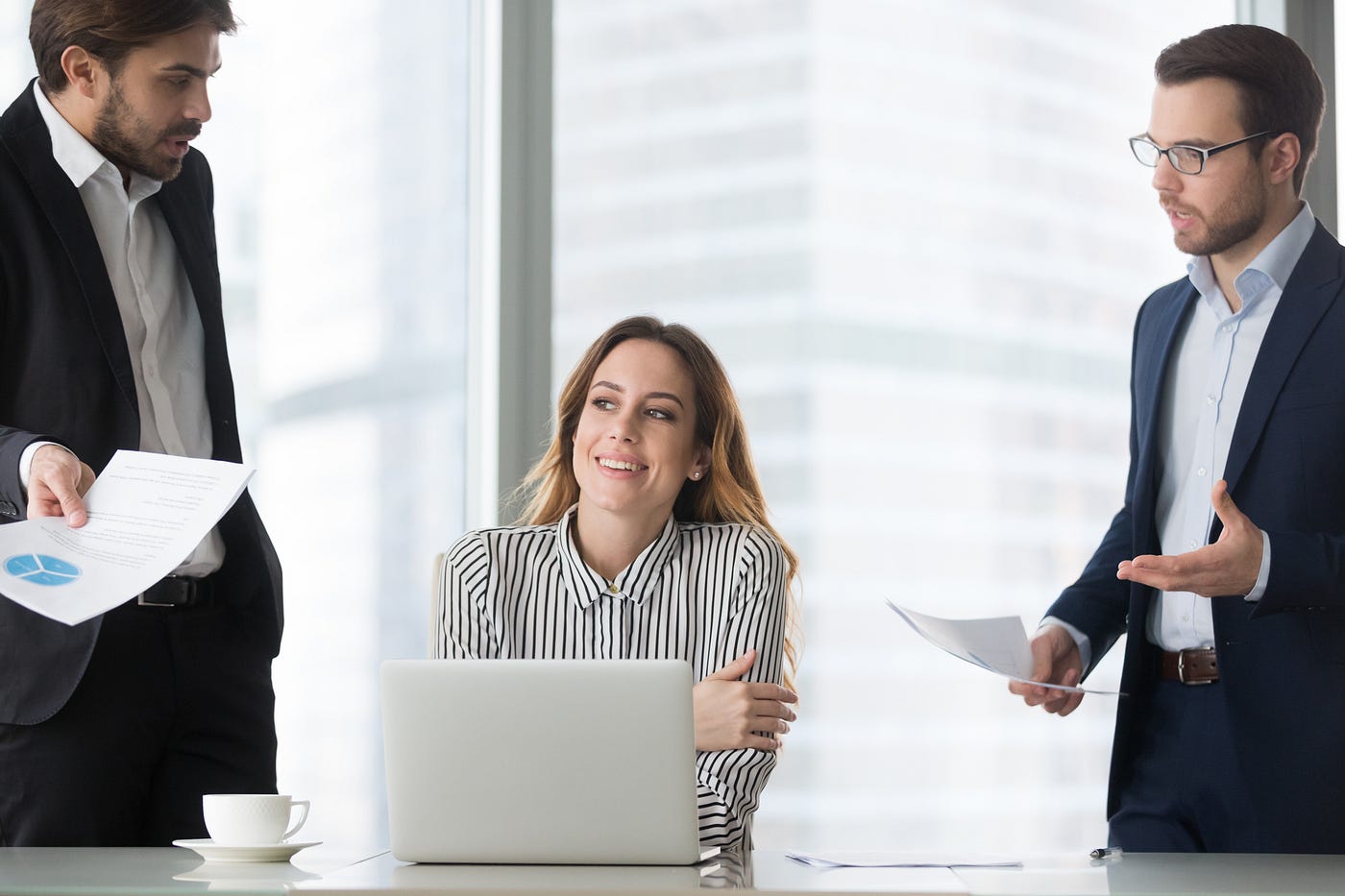 A businesswoman smiles awkwardly and looks away as two of her colleagues are bothering her with questions at her desk.