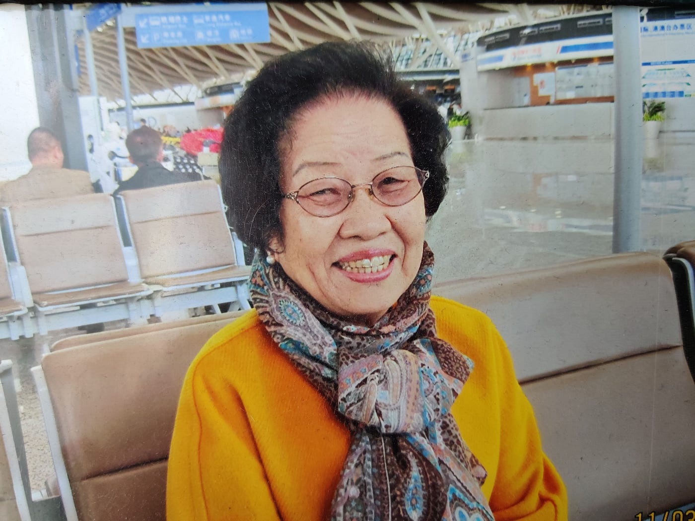 A smiling woman in a yellow sweater and scarf looks into the camera while seated in a transit terminal.