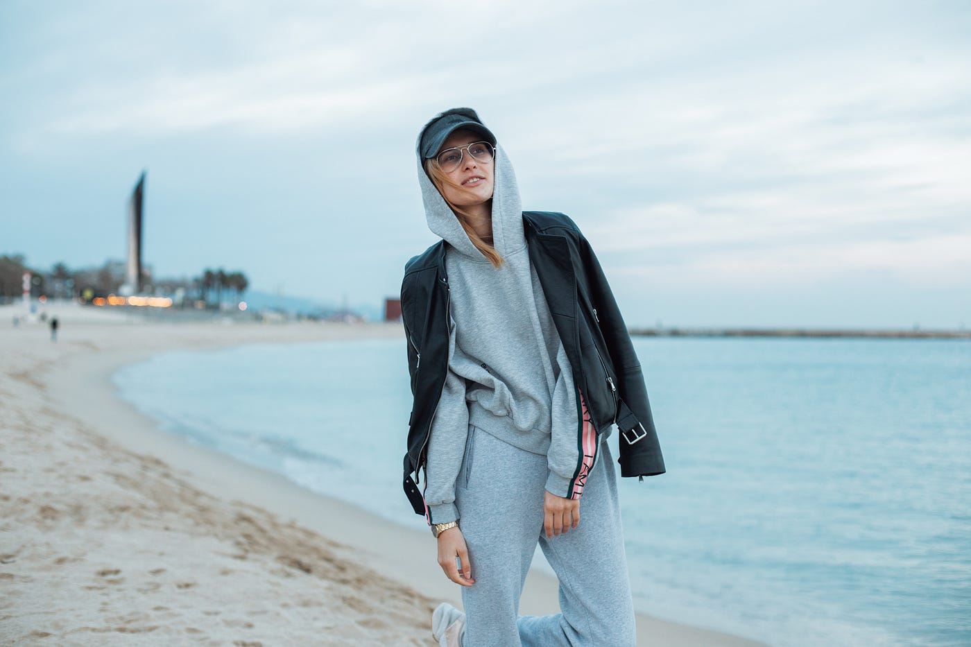 Woman in gray sweatsuit, baseball cap, and glasses with oversized coat hanging out on the beach on a blustery day