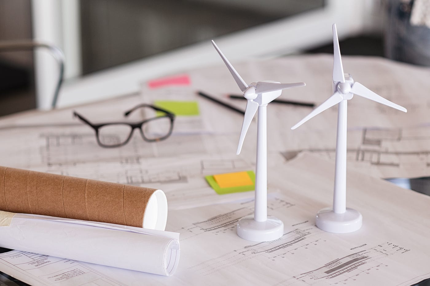 Two miniature white turbines sit on top of architecture plans for Passive House and net-zero construction next to a cardboard tube, a pair of glasses, and two pencils.