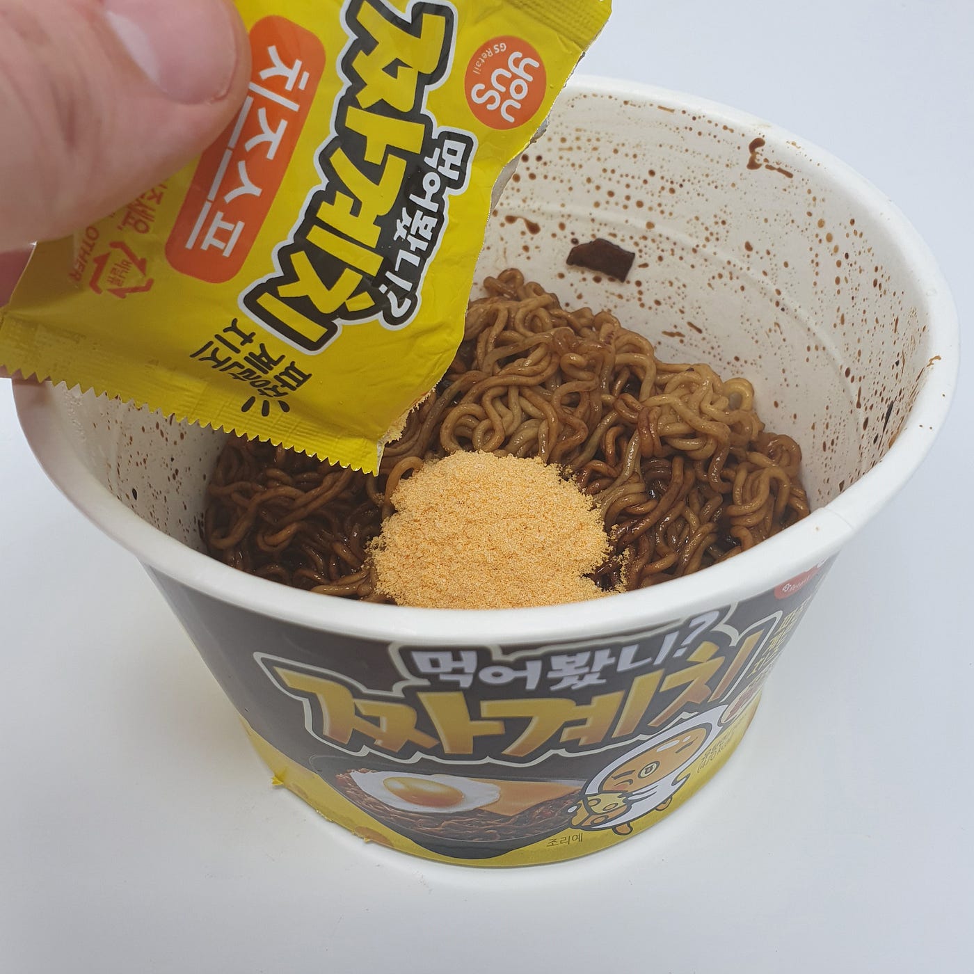Adding the powdered cheese soup packet to a cup of YouUs Jjagyechi Instant Noodles.