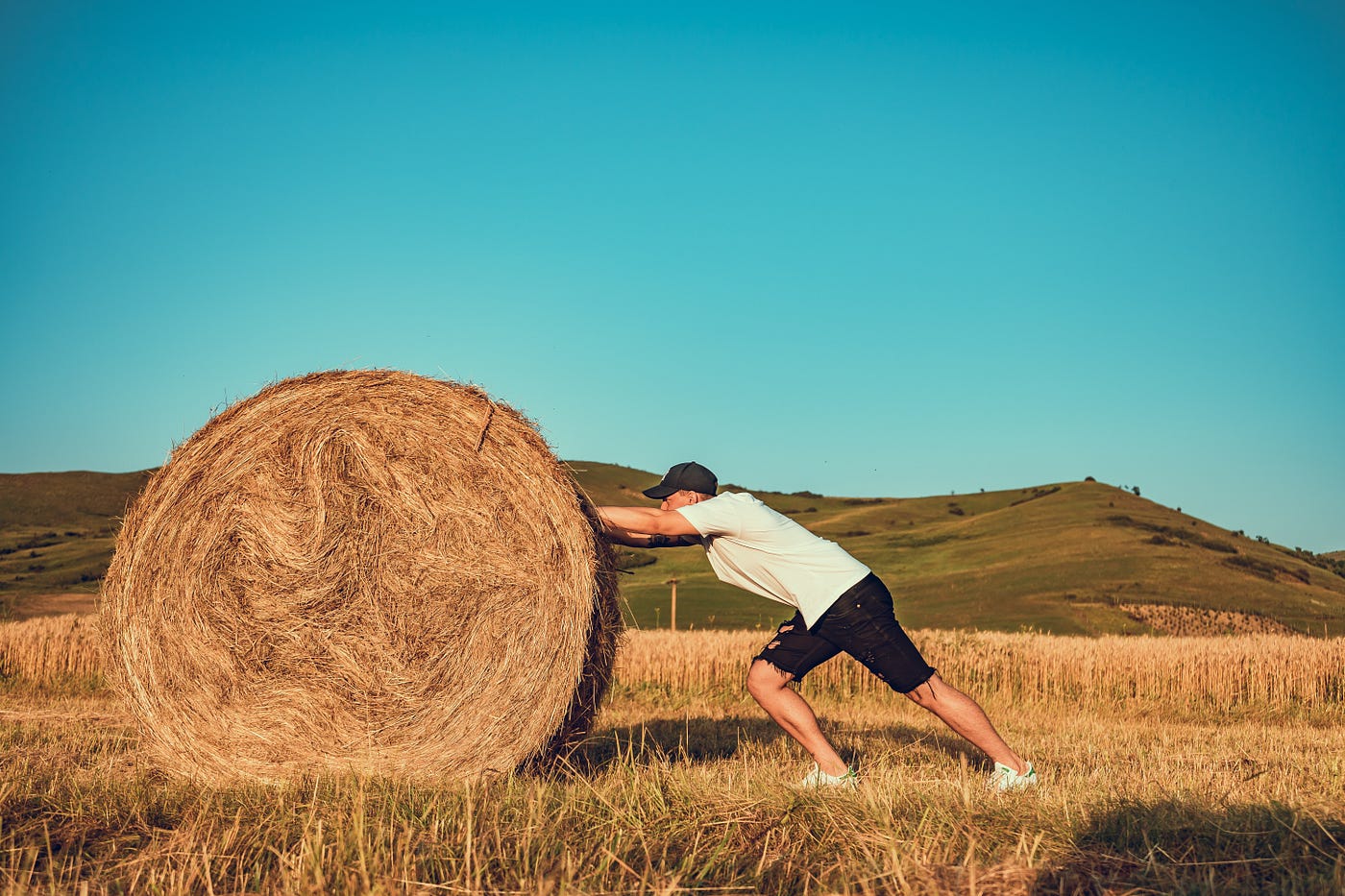 A man pushes a heavy looking hay bale in a golden field with hills and a blue sky in the background
