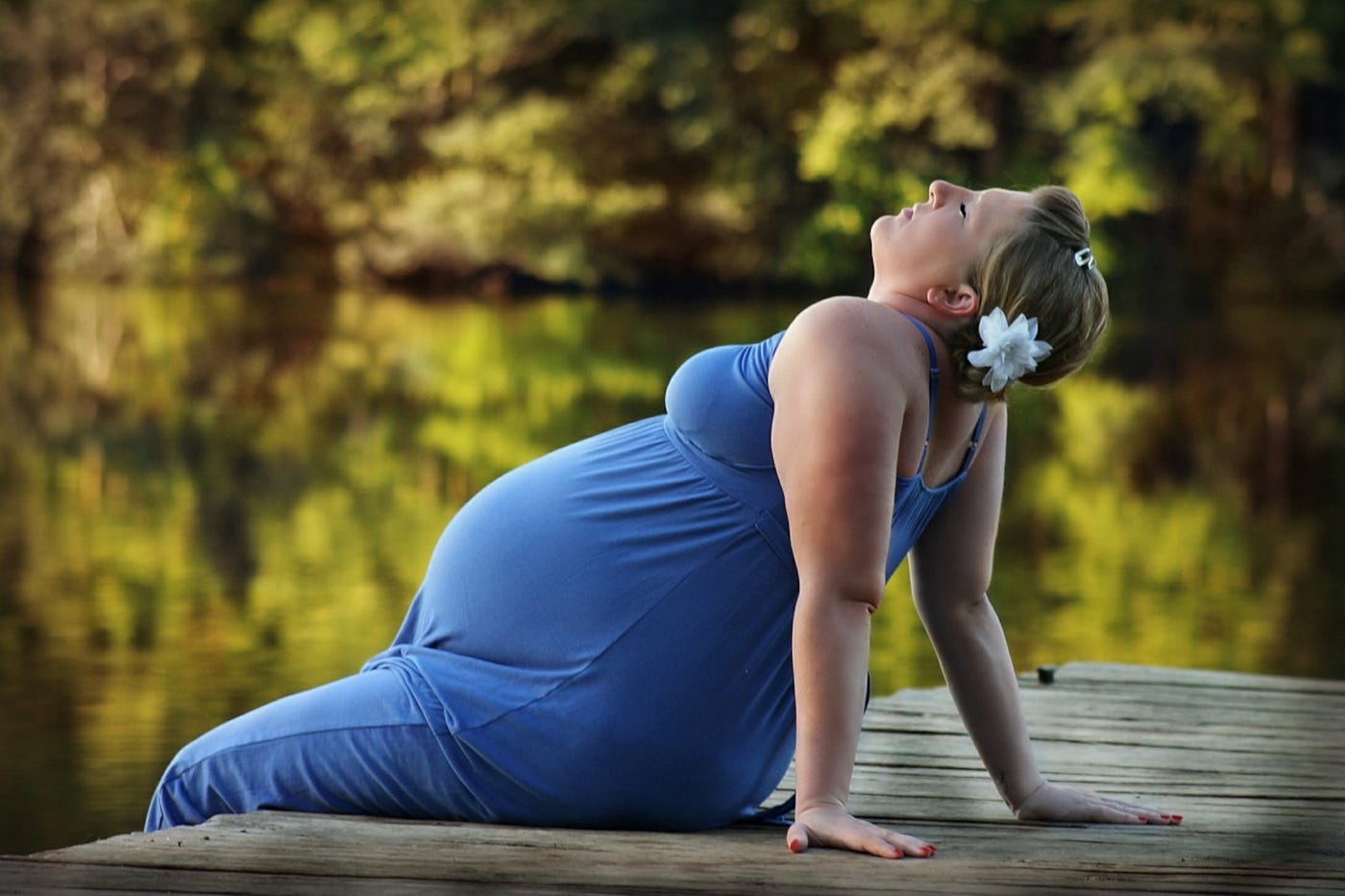 Color photo of a pregnant woman in a royal blue sleeveless dress with a flower in her hair. She’s relaxing on the edge of a boat dock, in the summer, enjoyinh  the moment.