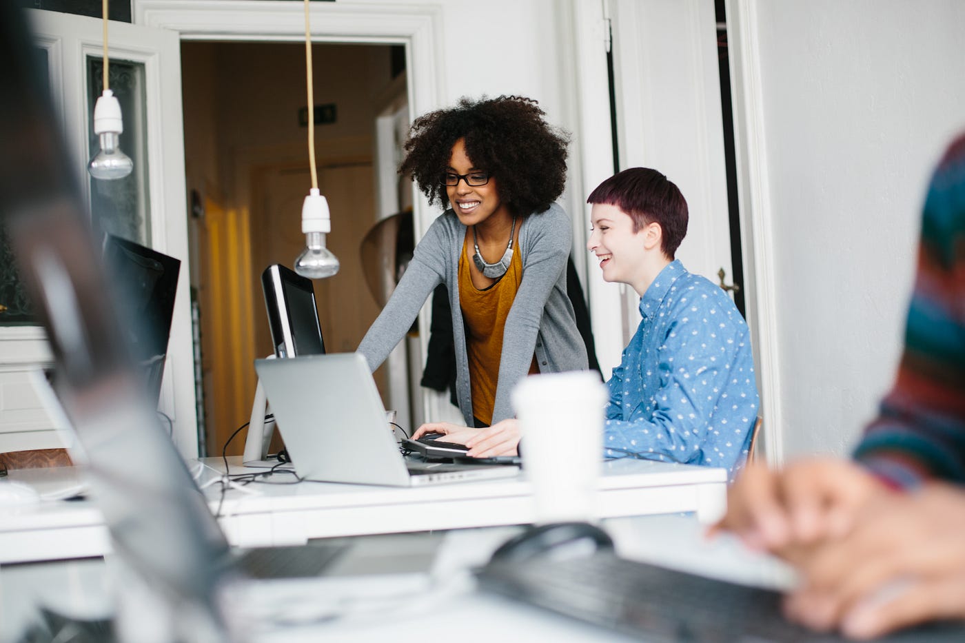 Two women entrepreneurs work together. One woman sits at her desk and the other is teaches her something off her laptop.