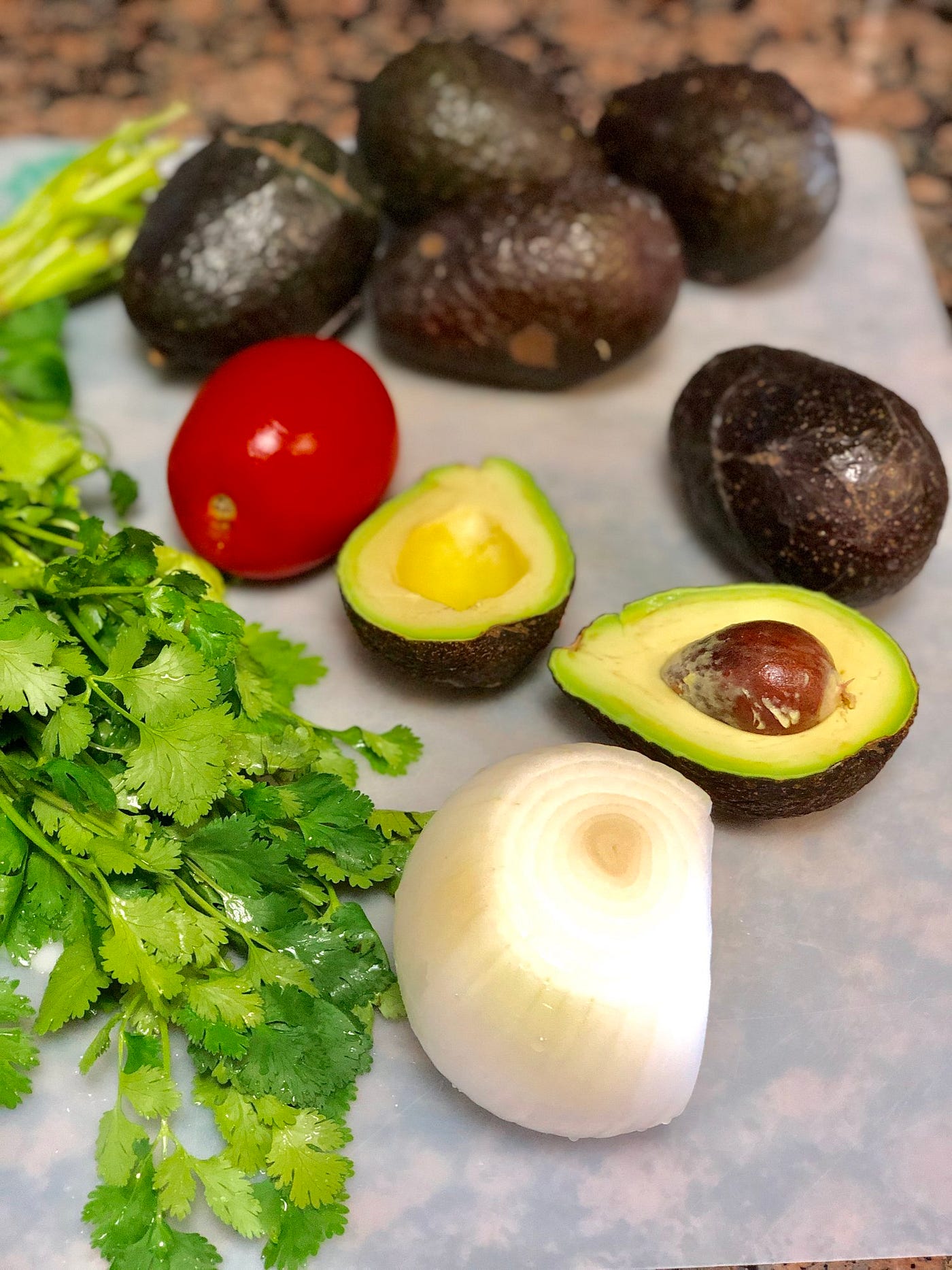 Ingredients to make guacamole. Cilantro, cut onion, avocado split in half, roma tomato on a plastic cutting board.