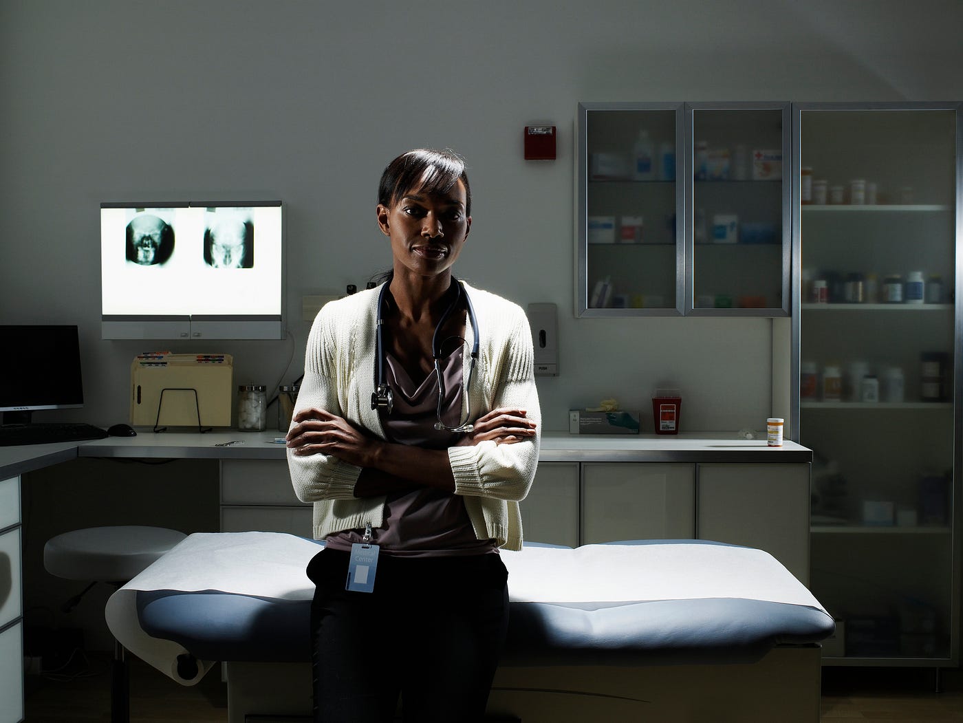 A photo of a Black female doctor sitting in an examination room, arms crossed.