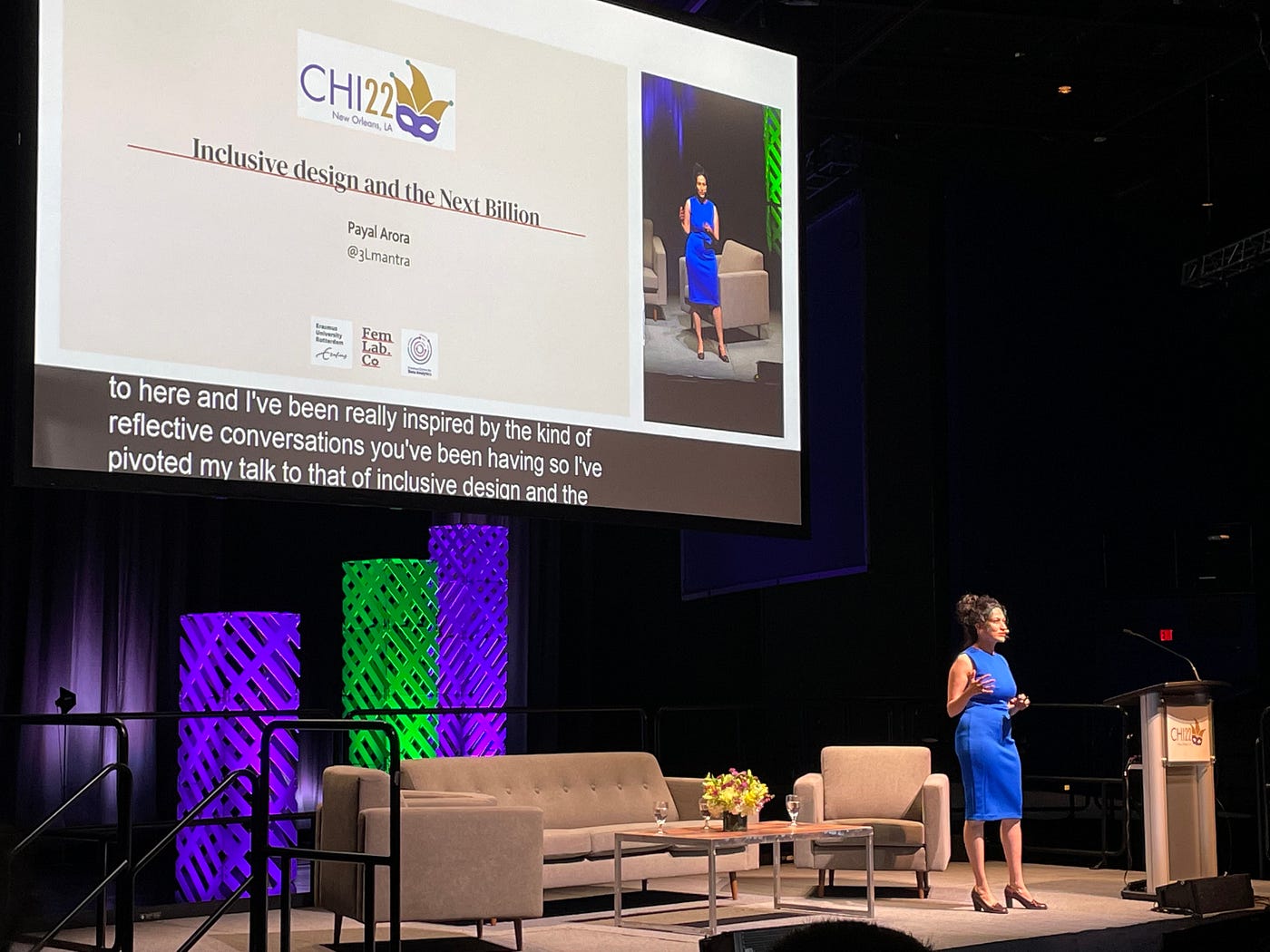 Payal in a blue dress on a stage with couches and a large projector screen with a slide reading “Inclusive Design for the Next Billion Users”