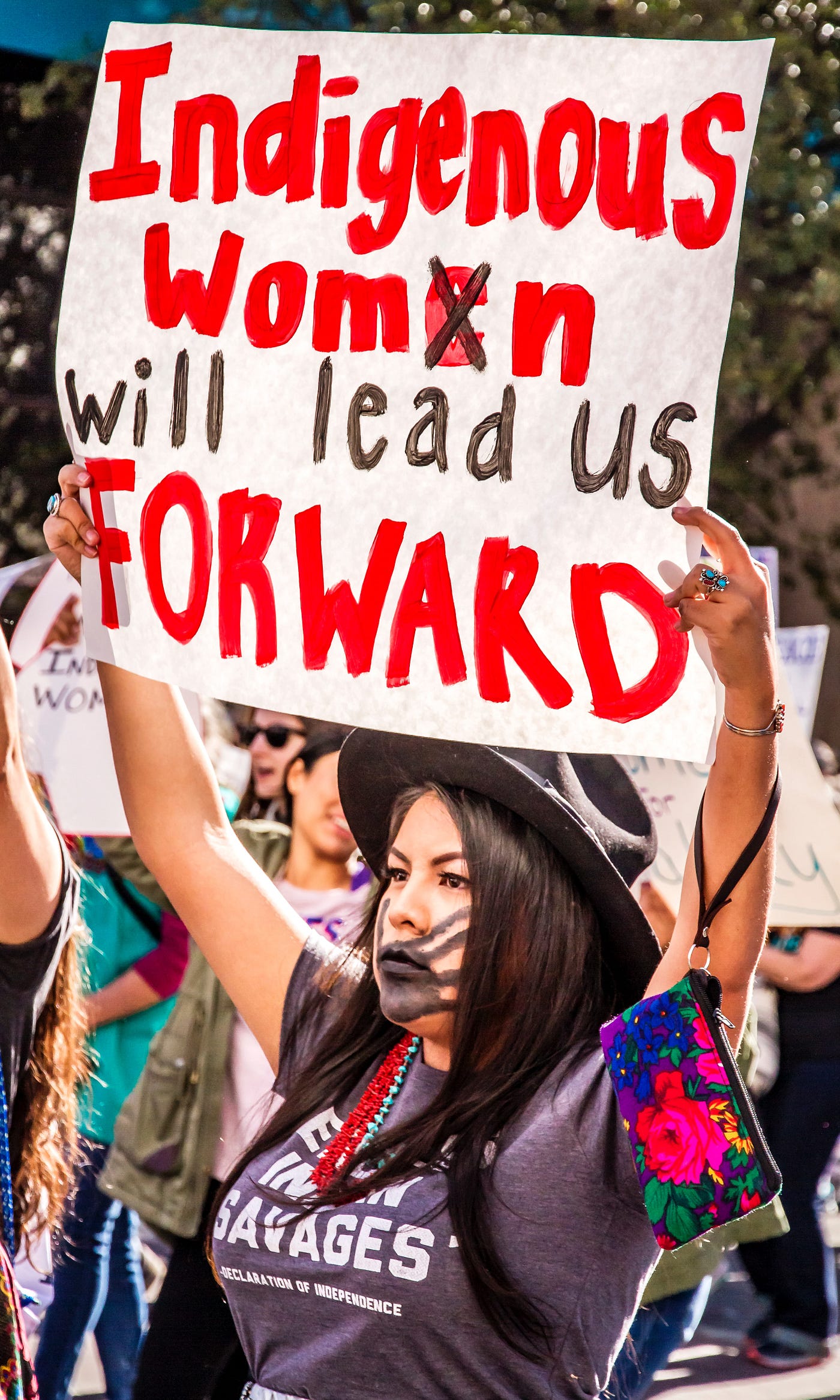 Protest. Indigenous woman holds sign, “Indigenous Womxn will lead us forward”. Black handprint painted over her mouth.