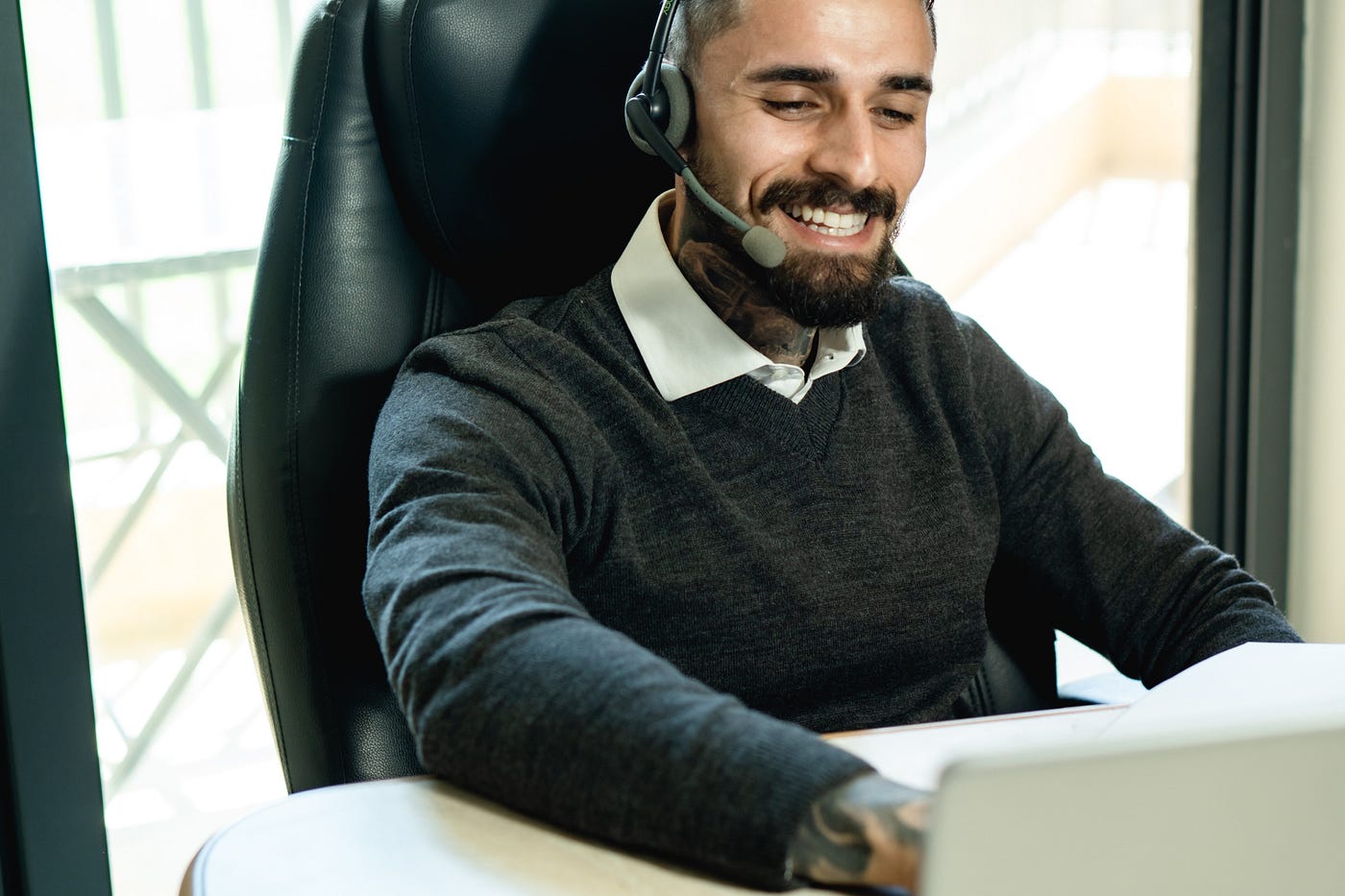 A man sitting at an office desk in business casual, smiling and having a headset on while looking at a laptop.