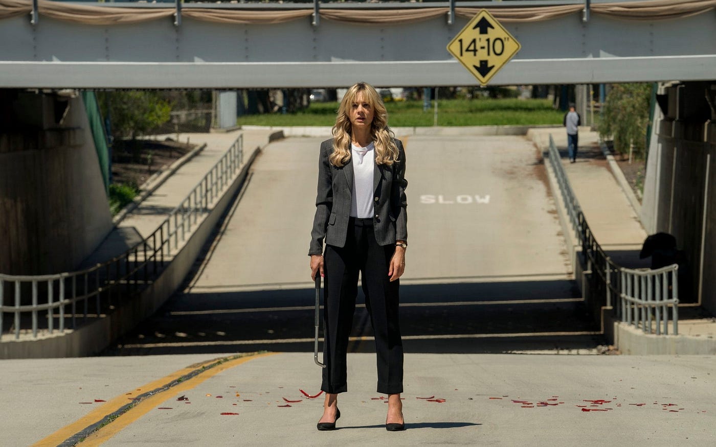 A blonde woman holding a crowbar, wearing a pantsuit in front of an underpass, with broken tail light shards on the ground.