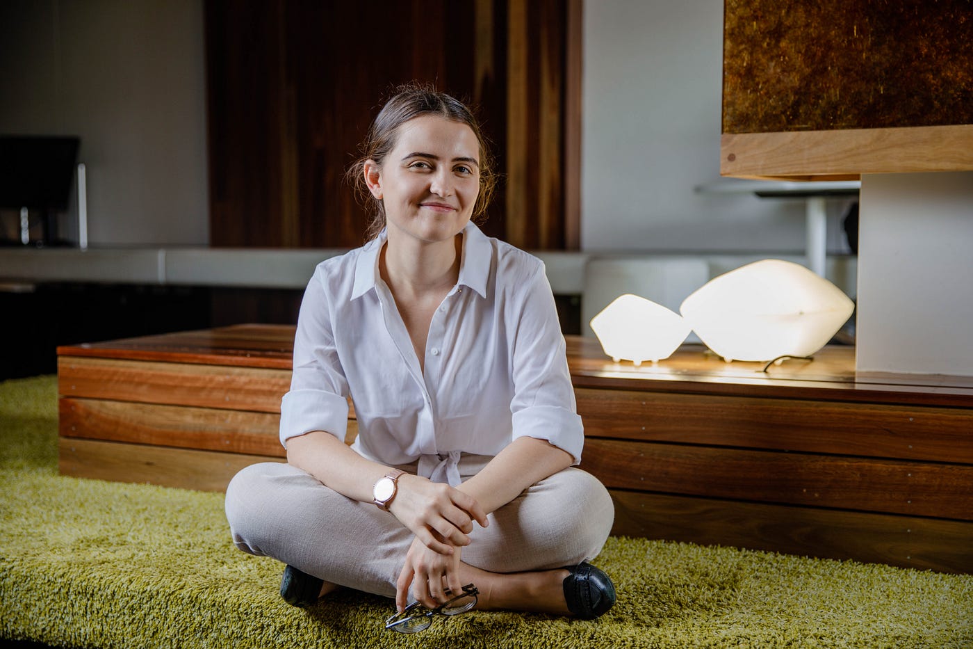 Ash Baxter, sitting cross-legged on the ground in a stylish room — plush green carpet, exposed wood and interestingly shaped lamps. She’s smiling warmly at the camera, wearing a button-up shirt, trousers and loafers.